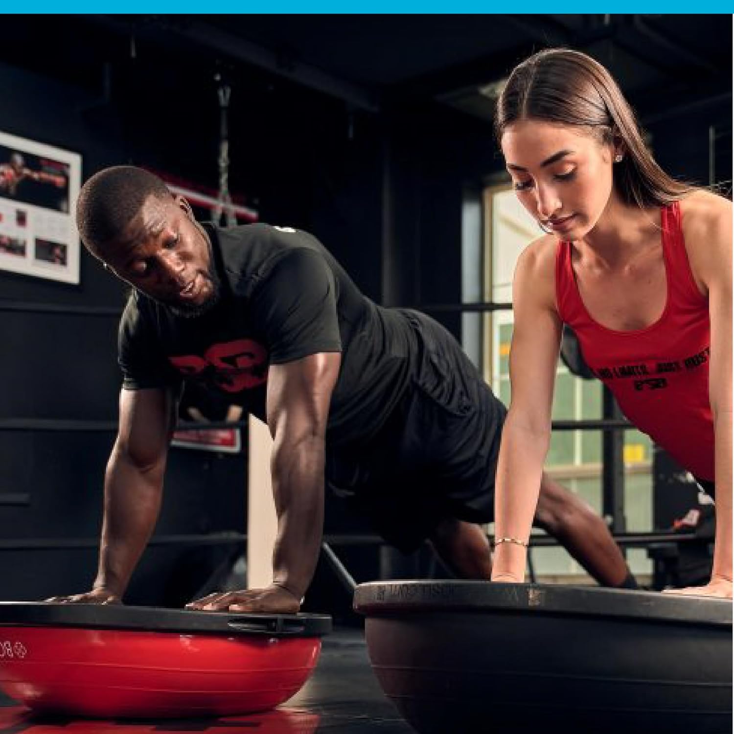 Couple Performing Push-Ups on BOSU Balance Trainers A muscular man in black athletic wear and a woman in a red tank top doing plank-style push-ups with their hands placed on red and blue BOSU Balance Trainers (dome side down, flat platform up) in a modern gym environment, focusing on core stability and upper body strength during an intense workout session.