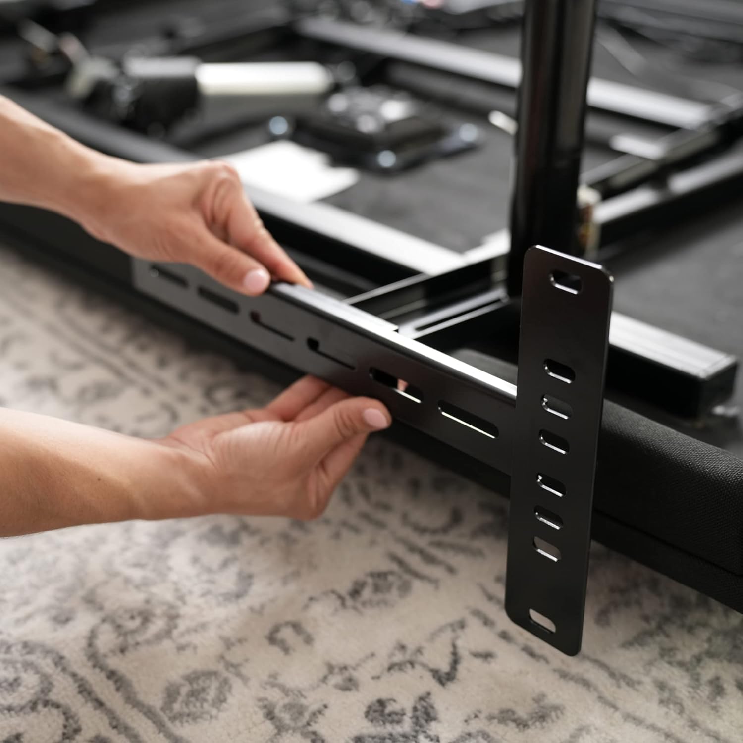 A person's hands holding a black T-shaped headboard bracket, demonstrating how its multiple slots allow for vertical adjustment along the bed frame.