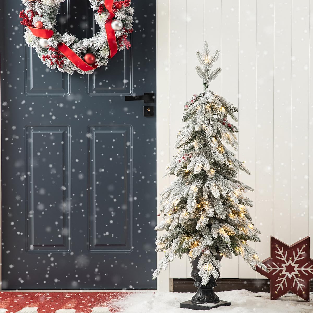 A single Glitzhome 4ft flocked Christmas tree with warm white lights and red berries, in an urn base, positioned next to a dark blue door with snow falling.