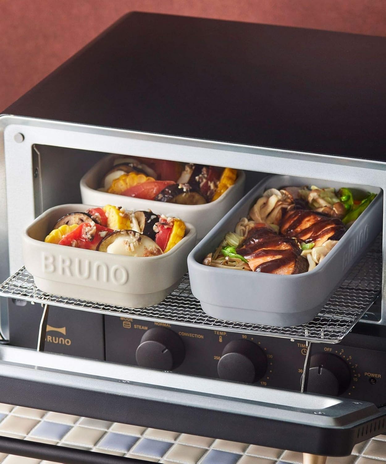 Two BRUNO ceramic toaster cookers placed on the rack inside a toaster oven, filled with food.