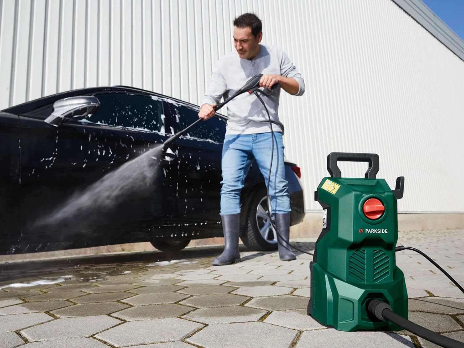 A person using the PARKSIDE PHD 110 E1 High-Pressure Cleaner to wash a black car on a paved driveway.