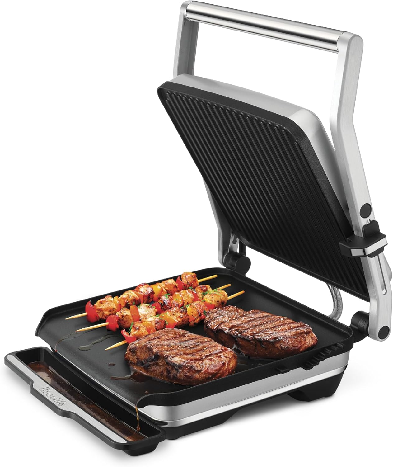 Breville AdjustaGrill & Sear open, showing grilled steaks and vegetable skewers on the cooking plates, with grease dripping into the tray.