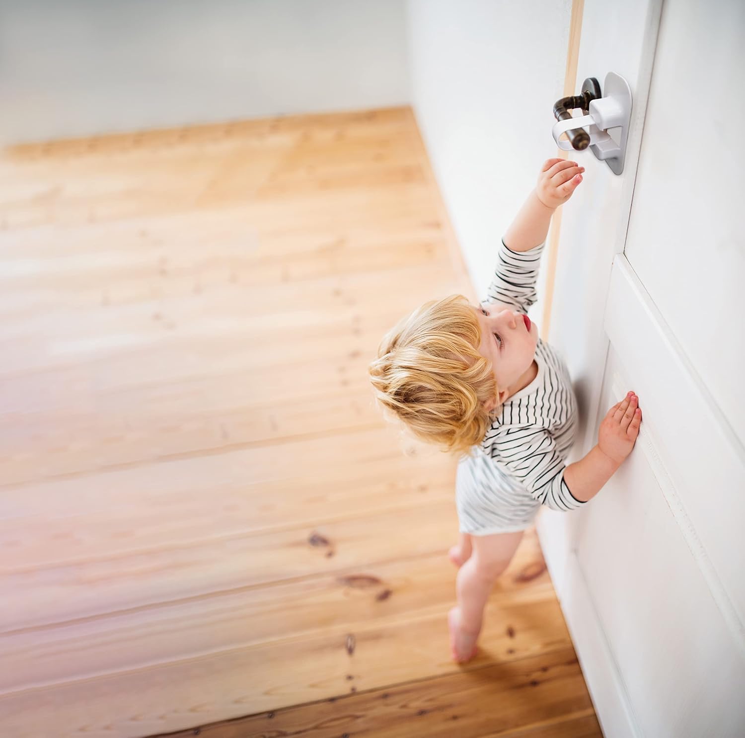 A young child reaching up towards a white door with the Dreambaby Adhesive Lever Door Handle Lock installed, demonstrating its purpose.