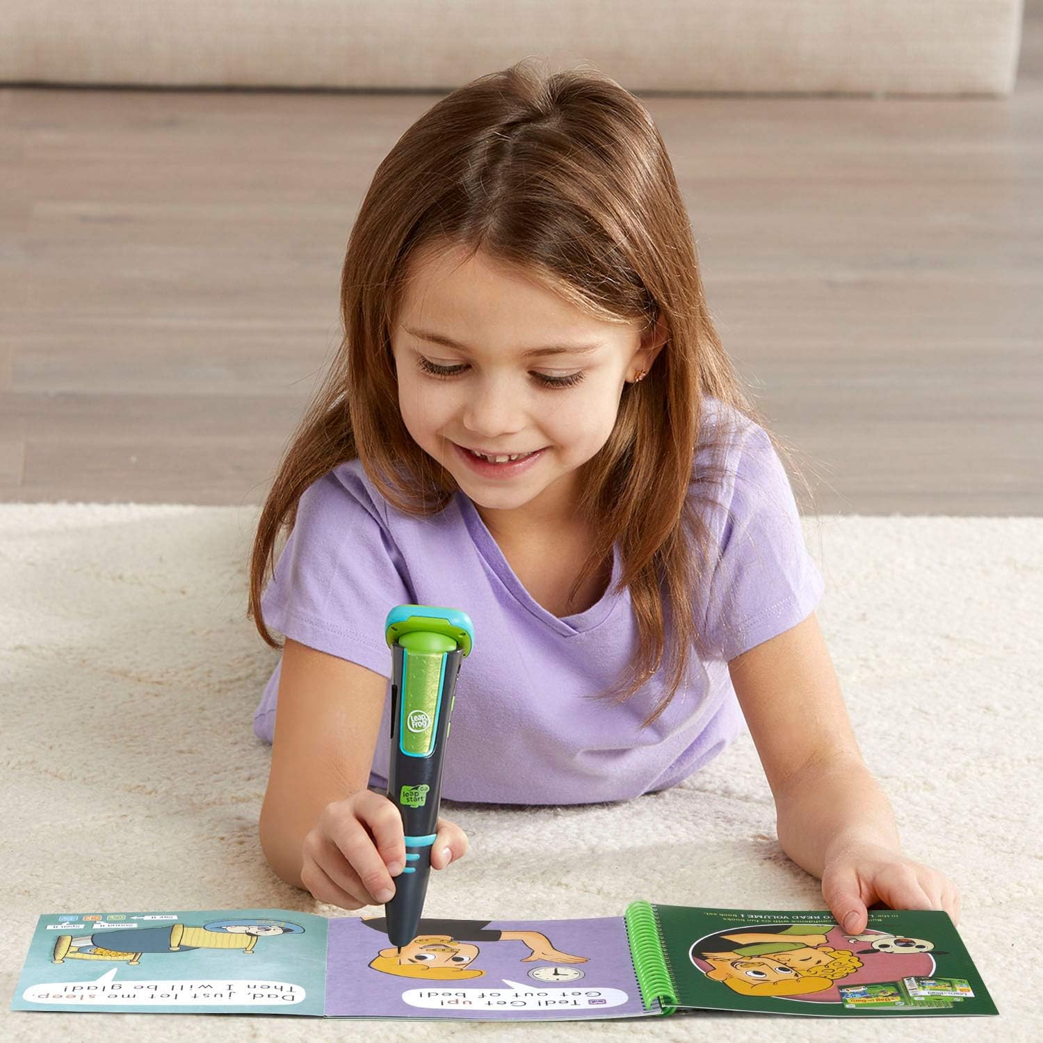 A young girl lying on a carpet, happily using the LeapFrog LeapStart Go stylus with an open activity book.