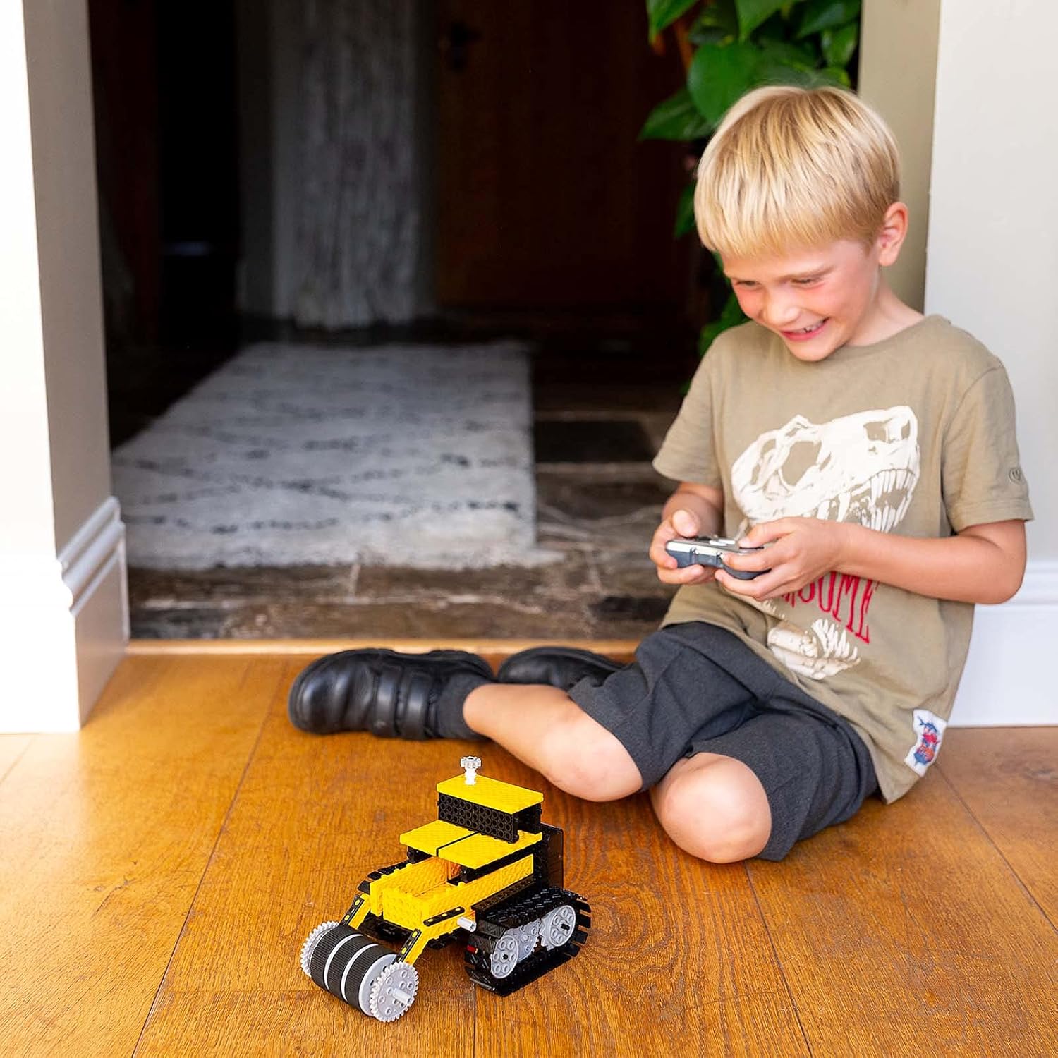 A child sitting on the floor, smiling and operating a remote-controlled construction vehicle (bulldozer) from the Think Gizmos set.