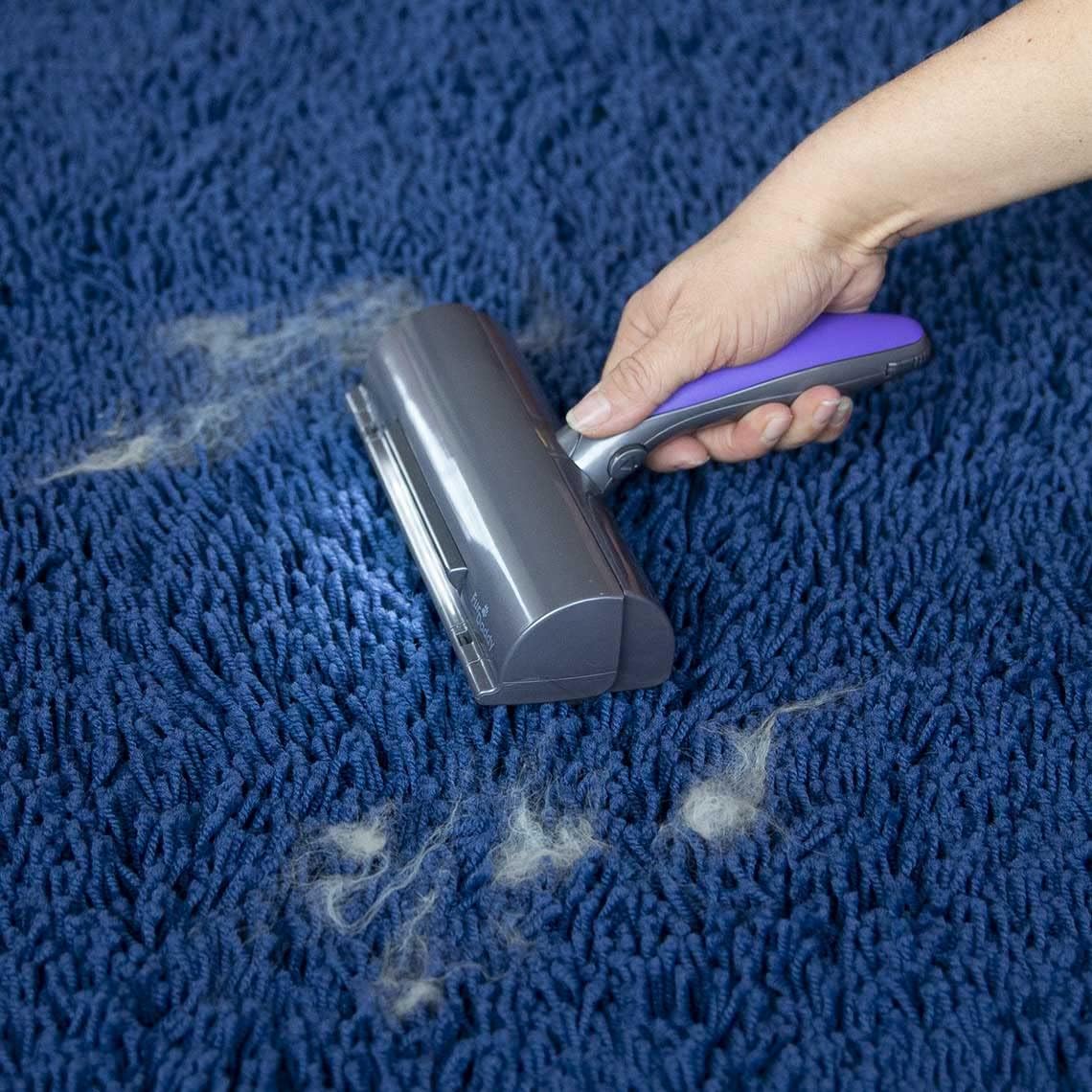 Hand using the Mediashop Fur Daddy Deluxe on a blue carpet, showing collected pet hair