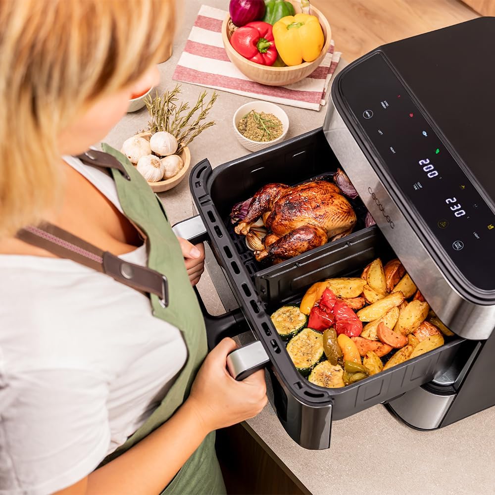 A person removing two separate compartments of cooked food (a whole chicken and roasted vegetables) from the Cecotec Cecofry FoodCourt 18000 air fryer, demonstrating the divider feature.
