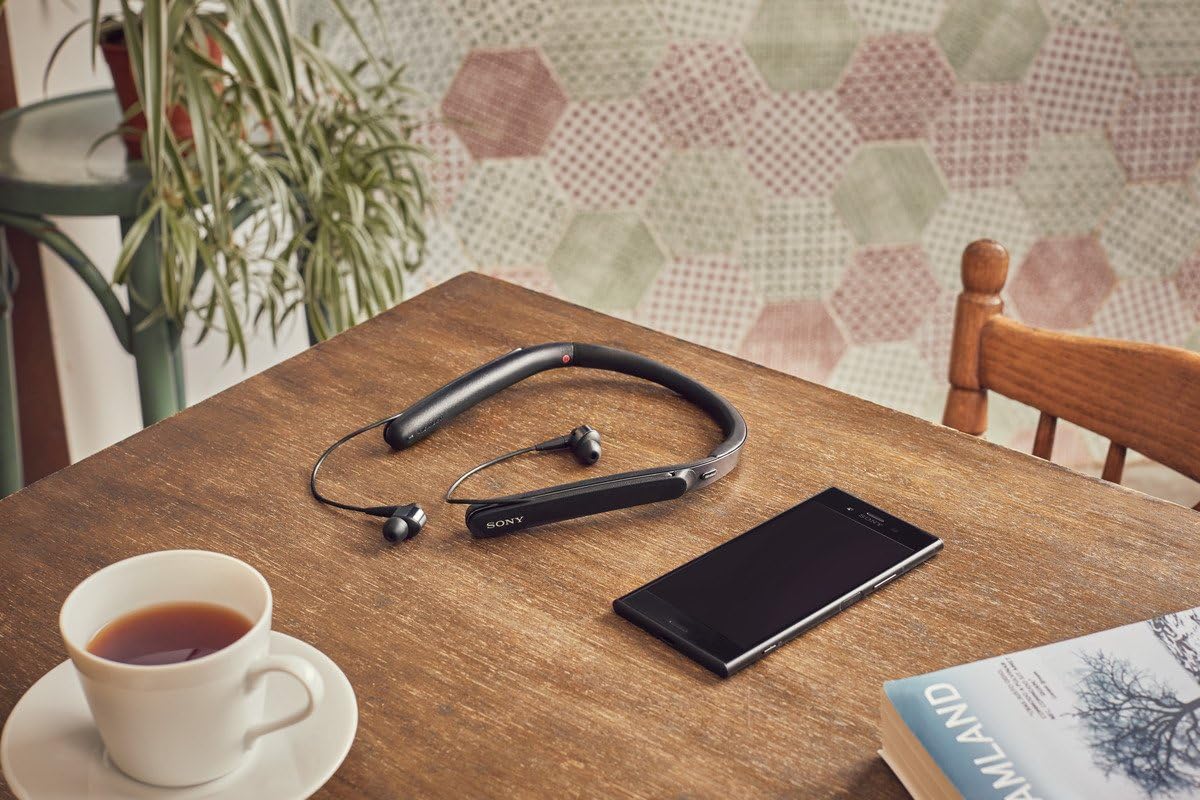 Sony WI-1000X headphones resting on a wooden table next to a smartphone, a cup of tea, and a book, illustrating everyday use.