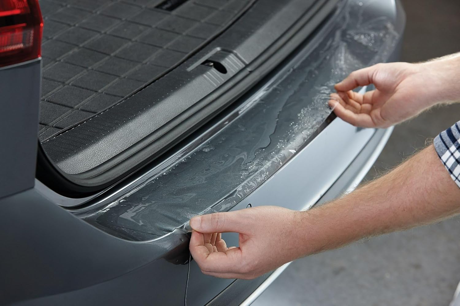 Hands using a squeegee to smooth out a clear protective film applied to a car's trunk ledge.