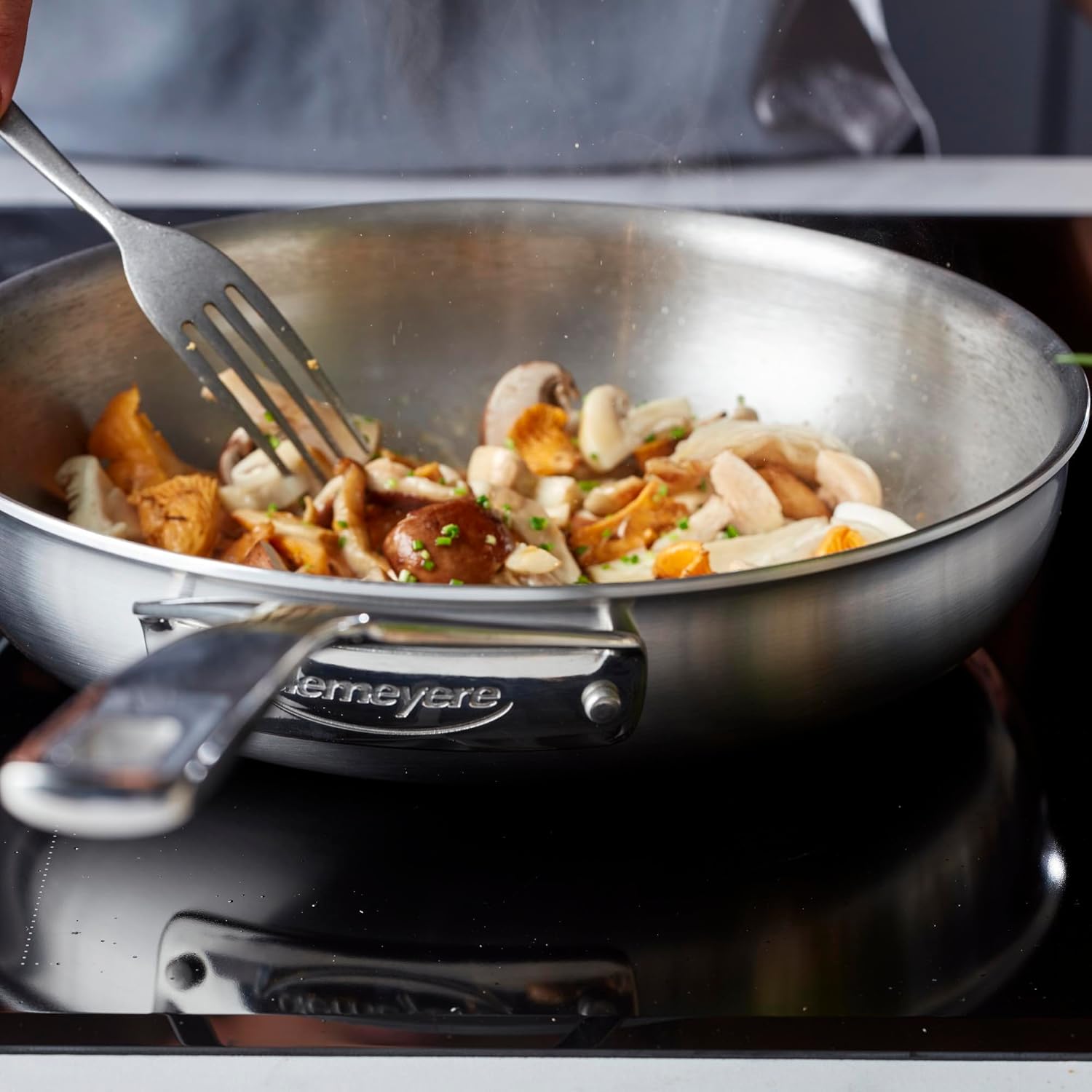 A chef cooking mushrooms and other ingredients in a Demeyere stainless steel fry pan on an induction cooktop.
