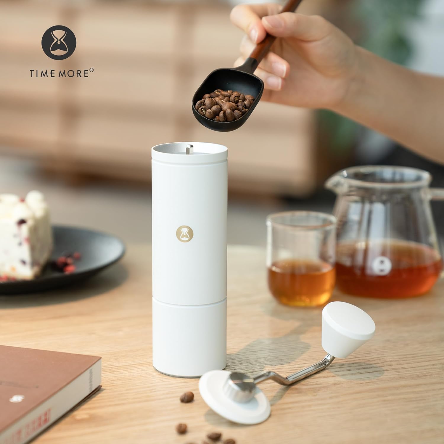 A hand pouring coffee beans from a scoop into the top opening of the TIMEMORE C5 hand coffee grinder.