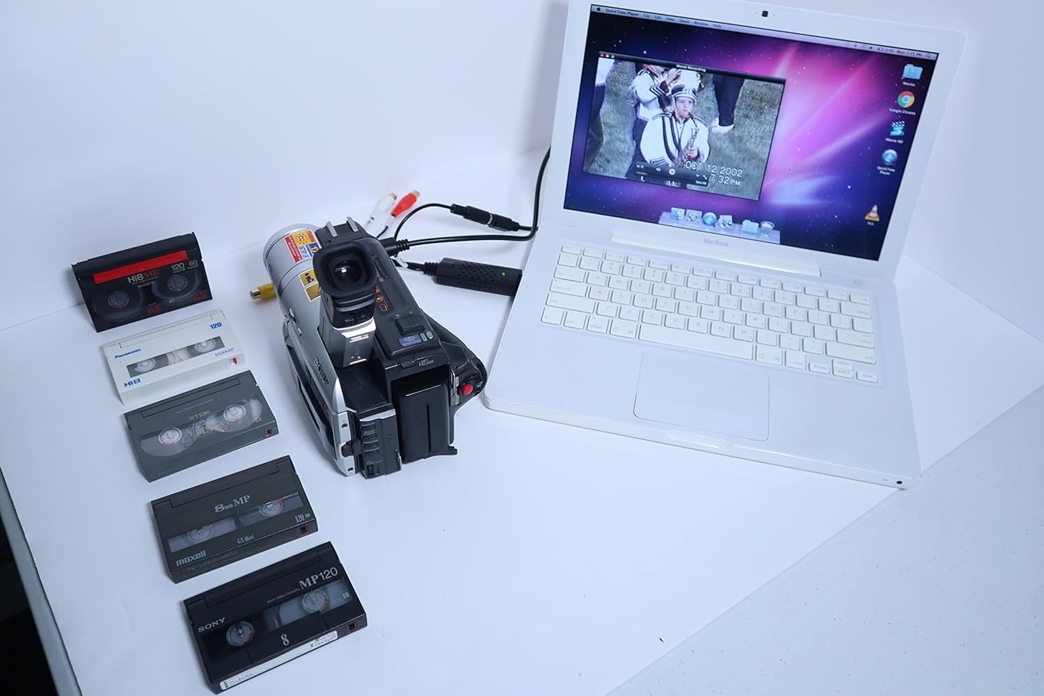 A Tech Collector camcorder connected to a white laptop via an AV to USB adapter, with various 8mm tapes laid out beside it. The laptop screen shows video playback.