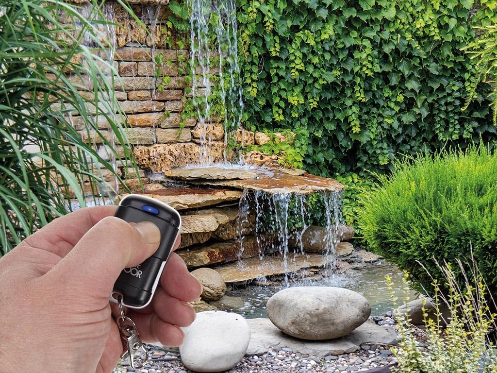 A hand holding the Unitronic remote control in an outdoor garden setting with a waterfall, illustrating its use for outdoor automation.