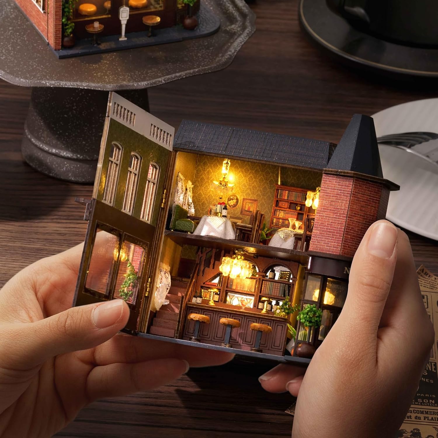 Hands carefully holding the partially assembled miniature Corner Restaurant, showing the intricate interior details and lighting.