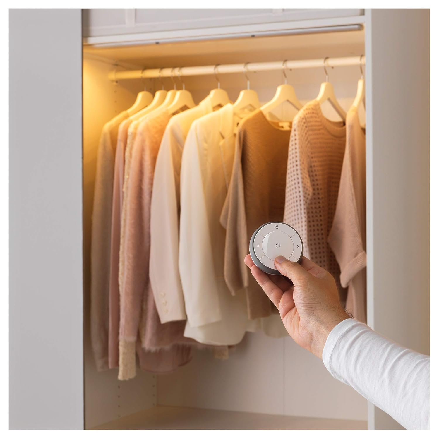 A hand holding an Ikea TRÅDFRI remote control, pointing towards an illuminated wardrobe with clothes, demonstrating light control.