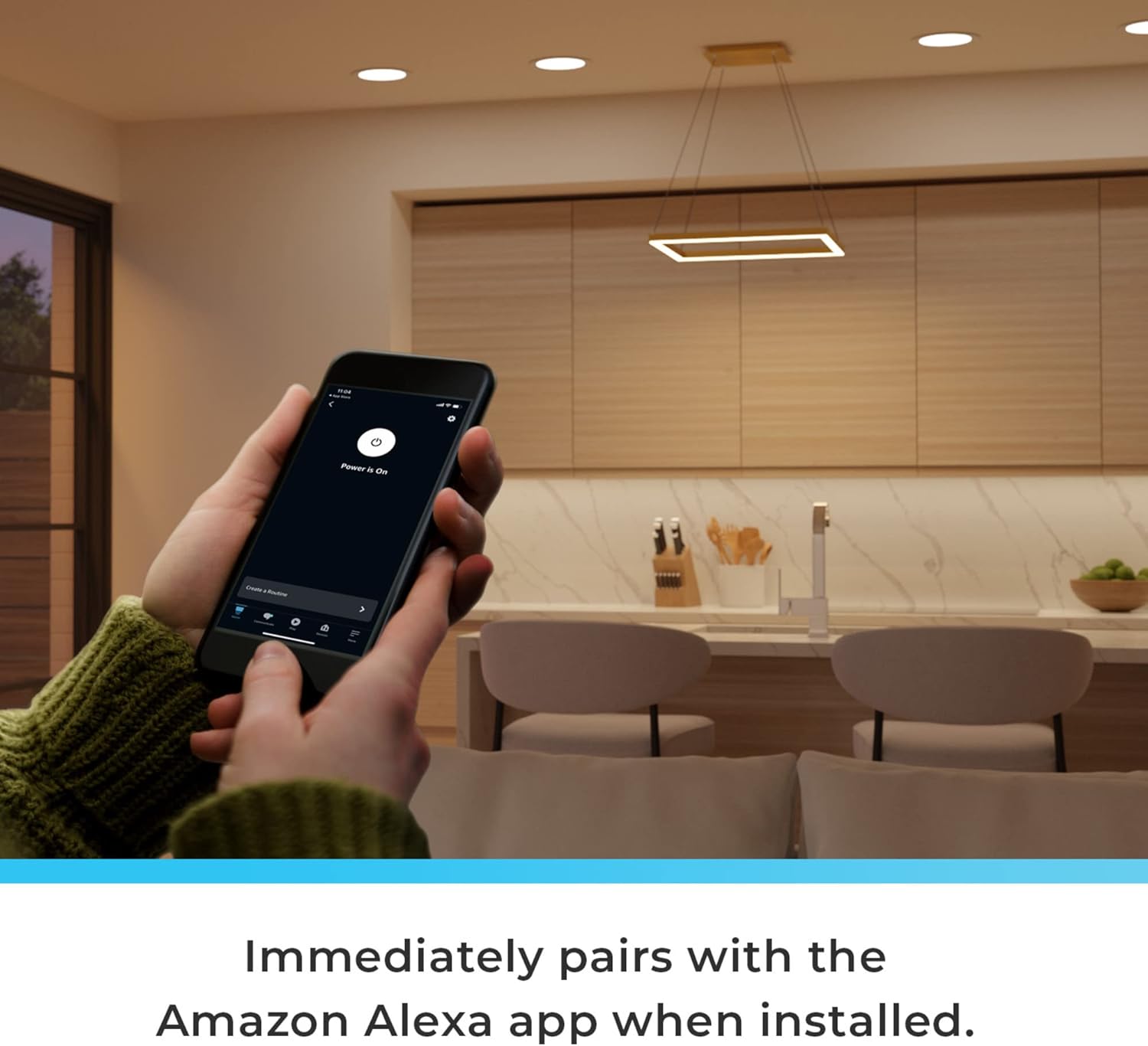 A person holding a smartphone with the Alexa app open, controlling the pendant light in a kitchen setting, with text 'Immediately pairs with the Amazon Alexa app when installed.'