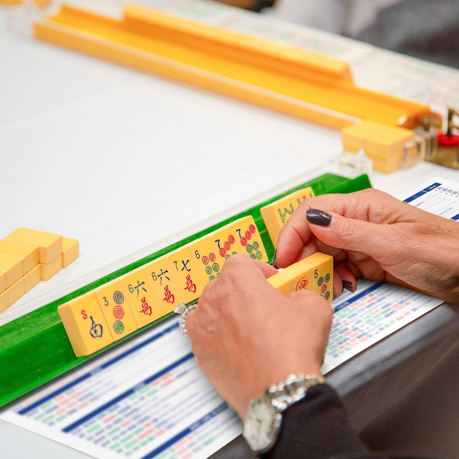 Close-up of a person's hands holding Mahjong tiles over an open AFSVTTS 2025 American Mahjong Card, showing the detailed rules.