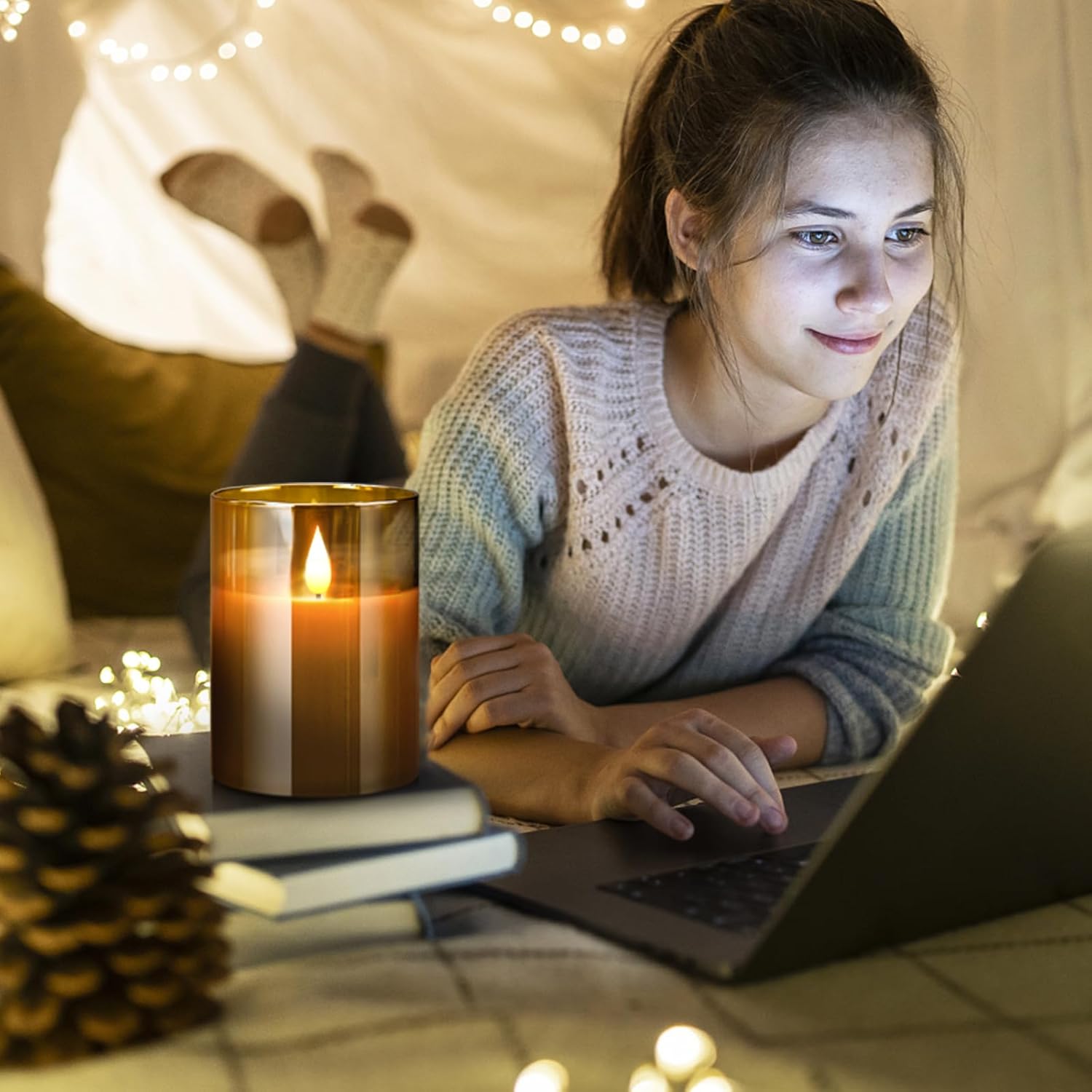 A flameless candle with a flickering light, placed on a table next to a laptop, with a person in the background. The scene is cozy and dimly lit.