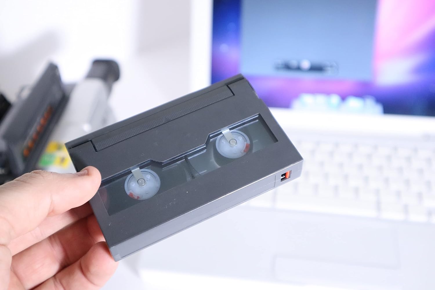 A hand holding an 8mm video cassette tape, ready to be inserted into a camcorder.