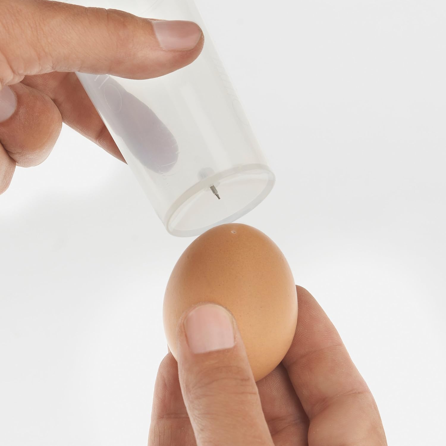 A hand holding an egg and using the egg piercer on the measuring cup to make a small hole in the egg.