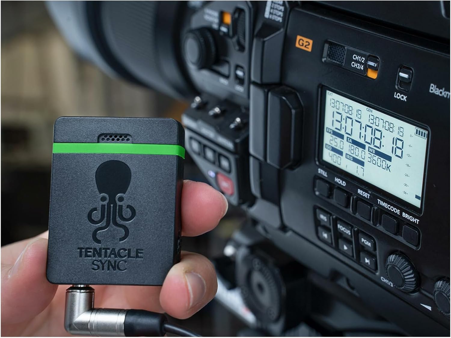 A hand holding a Tentacle Sync E MKII timecode generator next to a professional video camera displaying timecode on its screen.