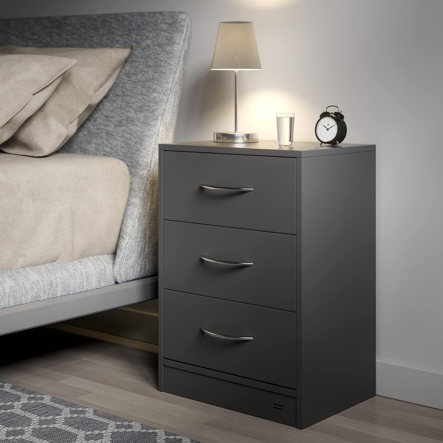 A single Casaria Eloise bedside table placed next to a bed in a bedroom setting, with a lamp and alarm clock on top.