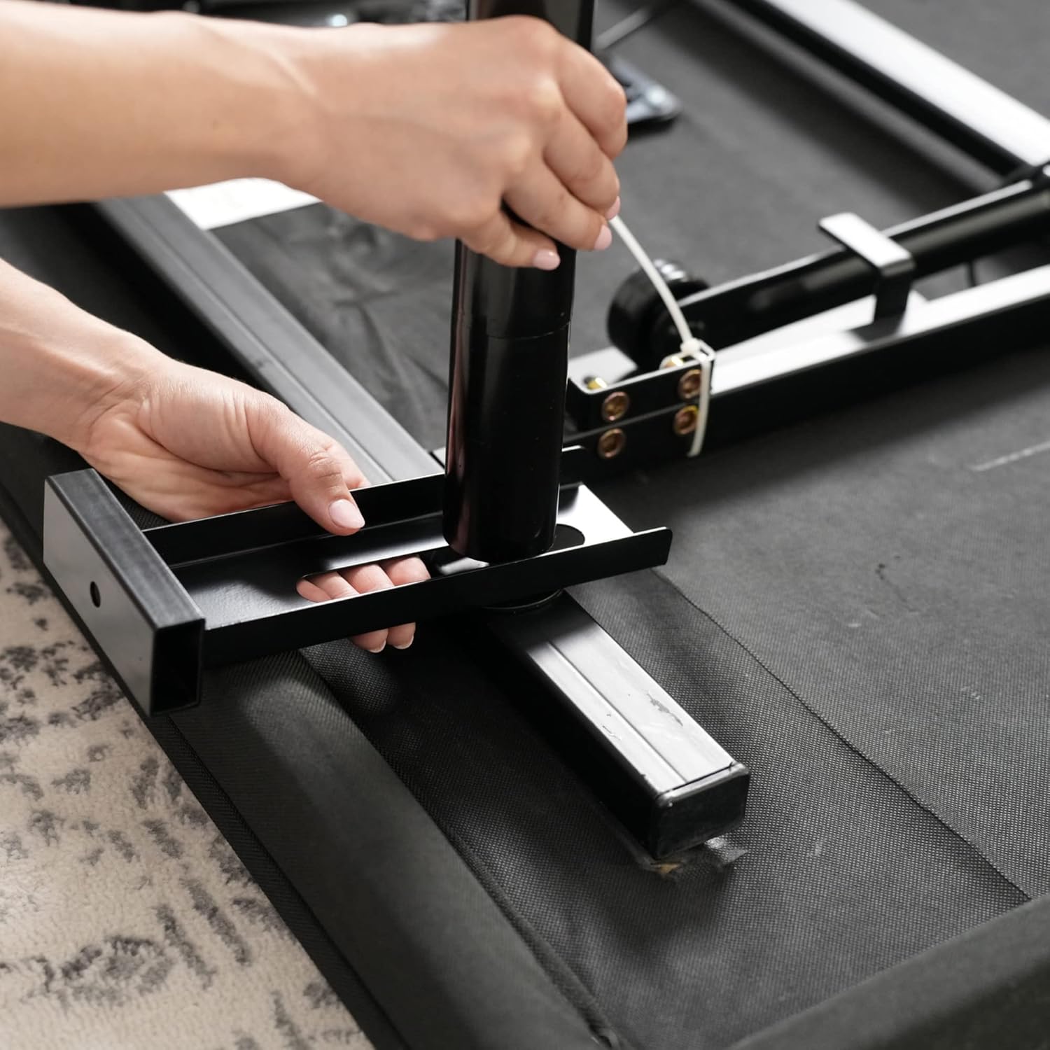 A person's hands attaching a black T-shaped headboard bracket to the side of an adjustable bed frame using a bolt and nut.