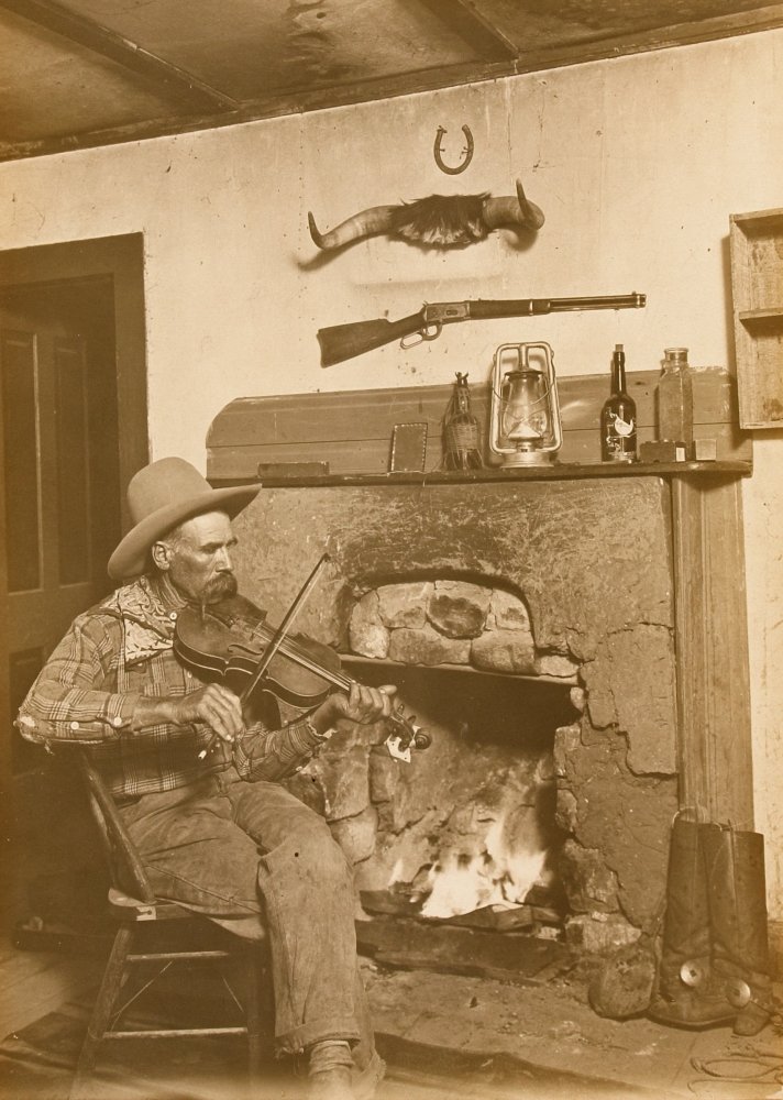 Erwin E Smith Three Block Cowboy New Mexico Cowboy Harry Patten Playing The Fiddle As He Sits Beside A Roaring Fireplace Cowboy Accoutrements (Boots Spurs Lantern Liquor Bottles Rifle Cow Horns And Lu