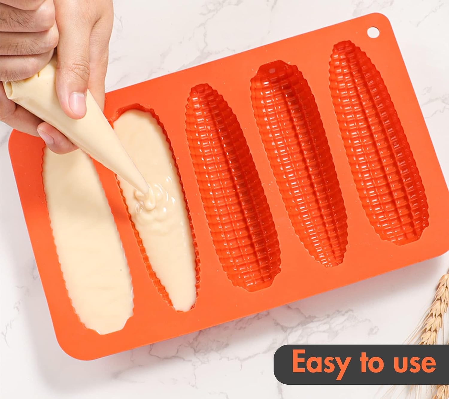 A hand using a piping bag to fill the corn-shaped cavities of an orange silicone baking pan with batter.