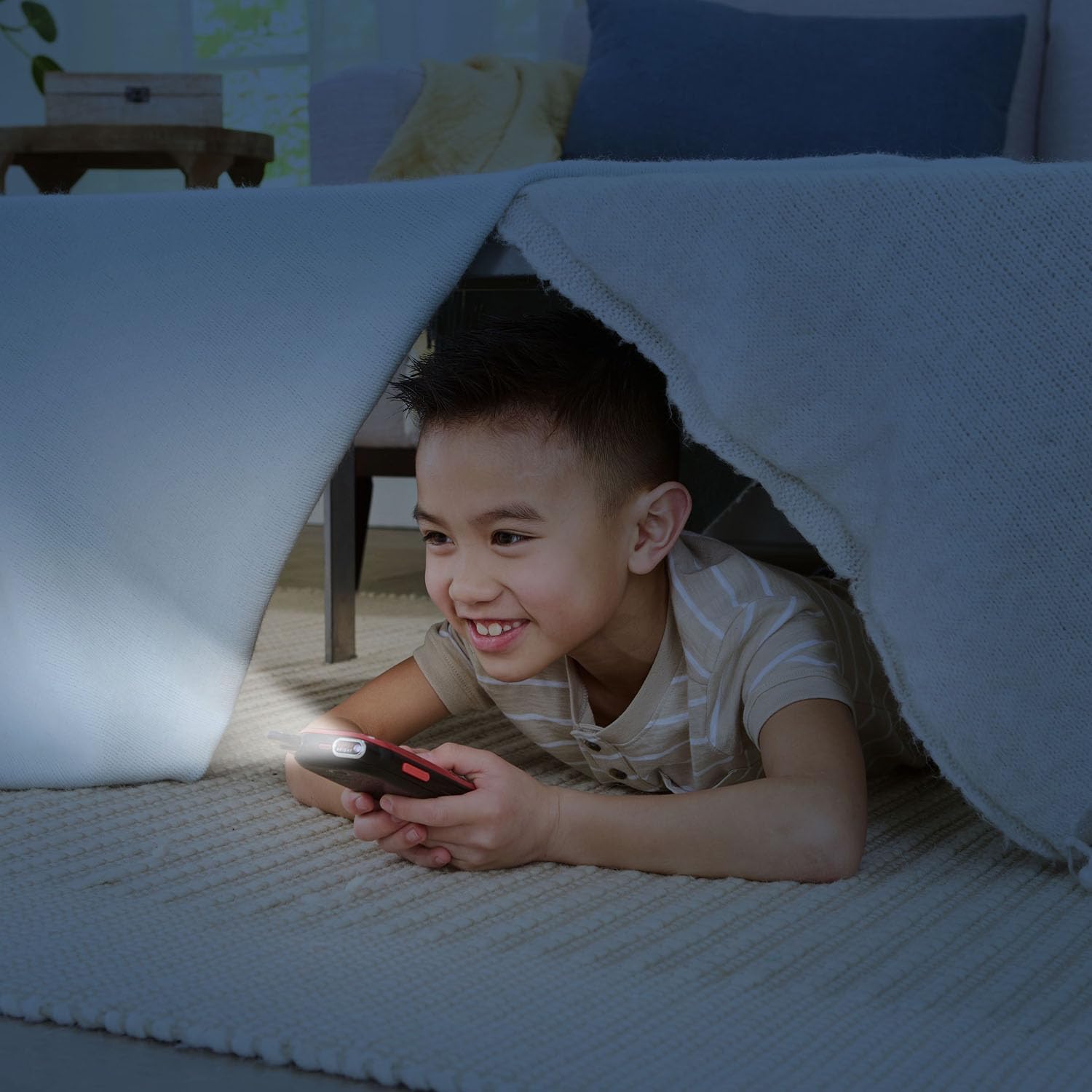 A young boy smiling while using the flashlight feature of a red VTech KidiGo Walkie Talkie DX under a blanket fort.