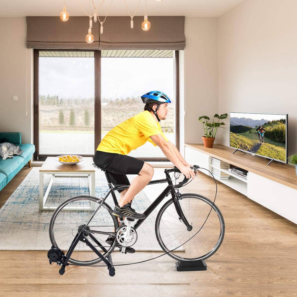 A man wearing a helmet and cycling attire is actively riding a black road bike mounted on the COSTWAY magnetic bicycle trainer indoors, facing a television displaying a cycling video. The trainer is set up in a modern living room.
