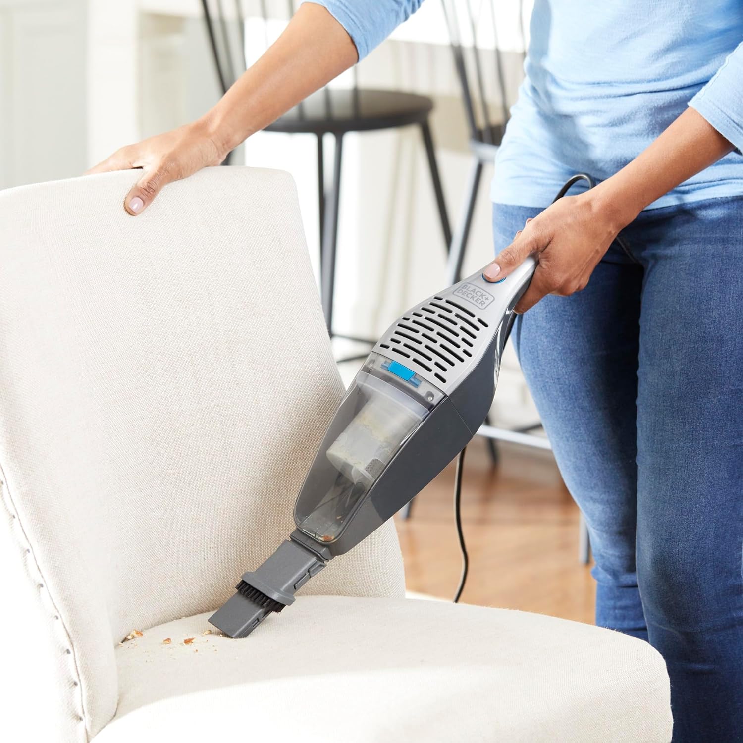 Woman using the BLACK+DECKER handheld vacuum with crevice tool on a wooden table.