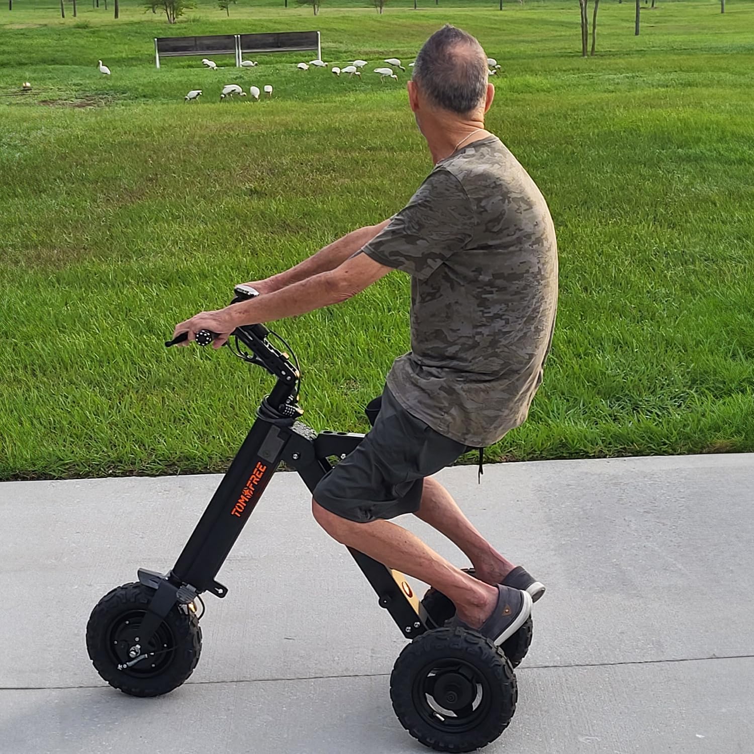 A person riding the Tomofree K7PRO 3-wheel electric scooter on a paved path with grass and trees in the background, demonstrating its use.