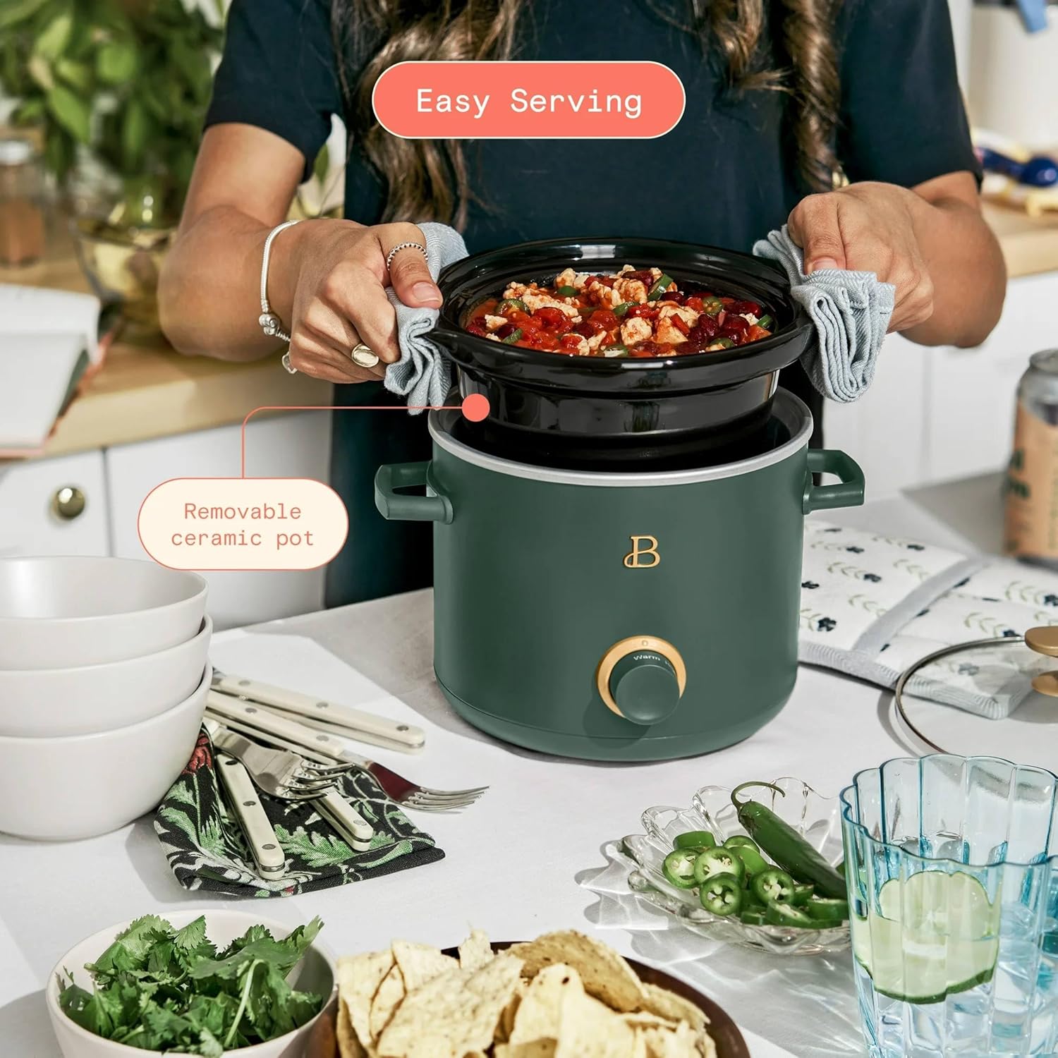 A person carefully lifting the removable ceramic pot from a Beautiful by Drew Barrymore slow cooker, demonstrating easy serving.