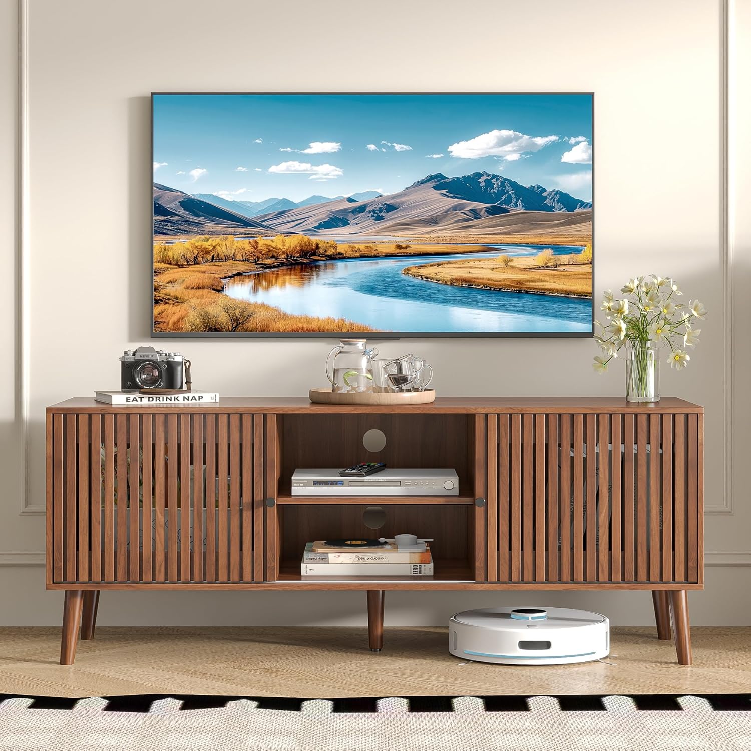 Oubayajia TV Stand in a living room setting with a television mounted above it. The stand is walnut-colored with slatted sliding doors. The central compartment is open, displaying a media player and books. A robot vacuum is visible underneath.