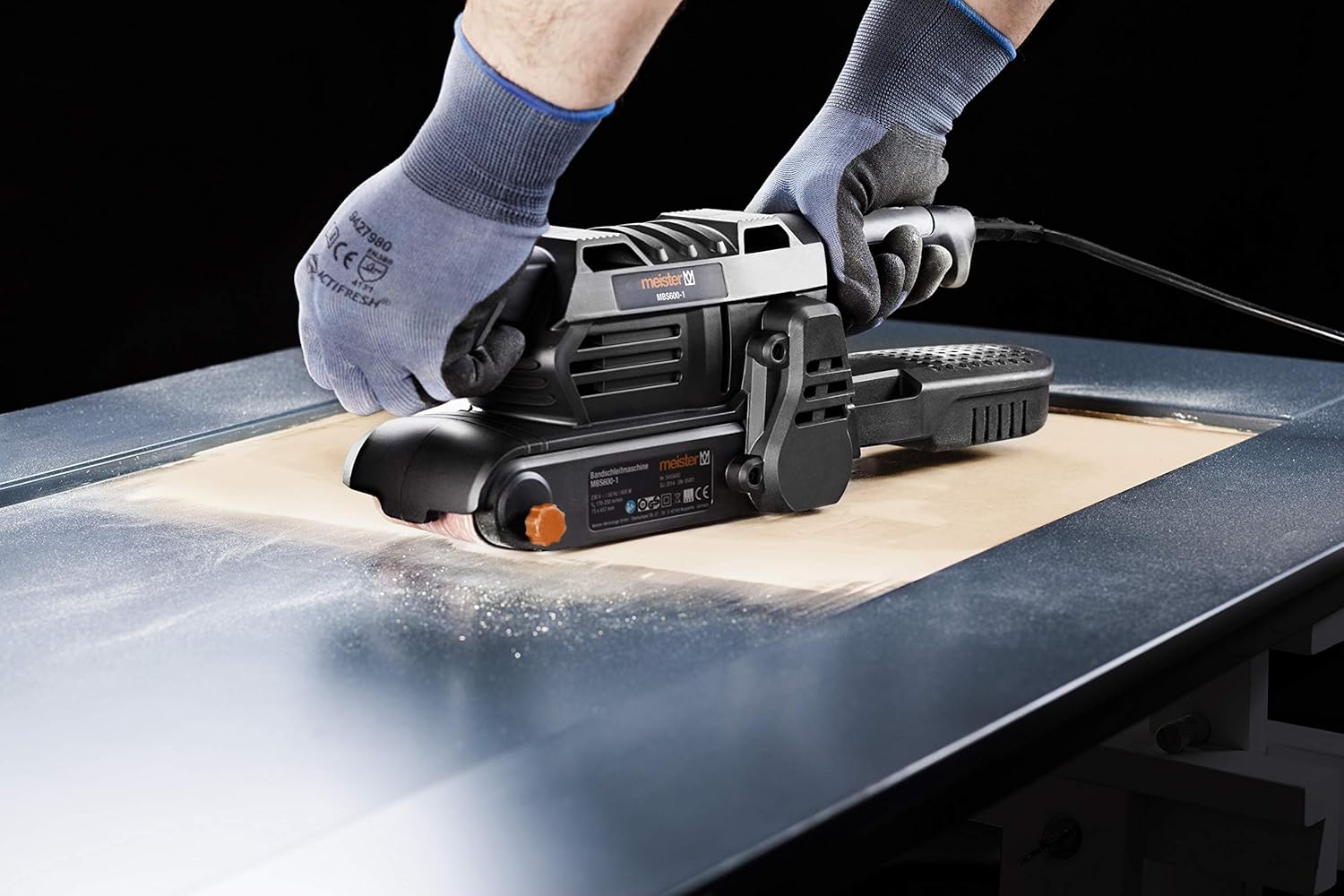 A person wearing gloves operating the Meister belt sander on a large flat surface, creating dust.