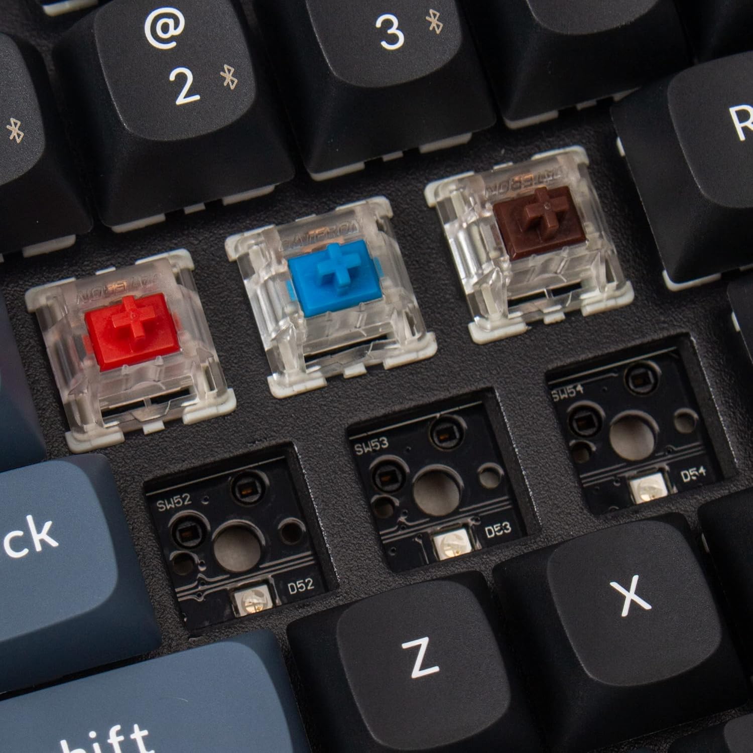 Close-up of the Keychron K8 Pro keyboard with several keycaps and switches removed, revealing the hot-swappable sockets and different colored switches.