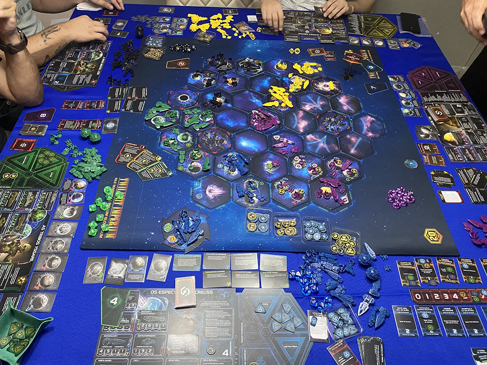 Vista aproximada de uma partida de Twilight Imperium 4ª Edição em um tapete azul, mostrando miniaturas de naves espaciais coloridas e planilhas de facção.