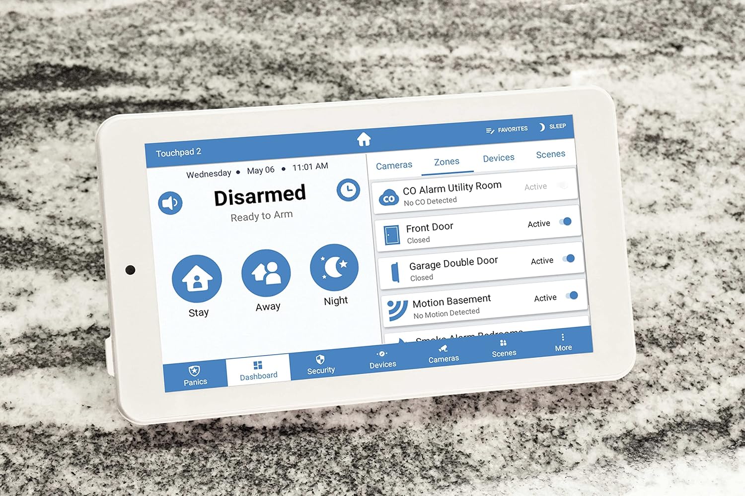 Alula Slimline Touchpad Pro resting on a countertop, displaying the main screen with tabs for Cameras, Zones, Devices, and Scenes, showing active zones like CO Alarm Utility Room and Front Door.