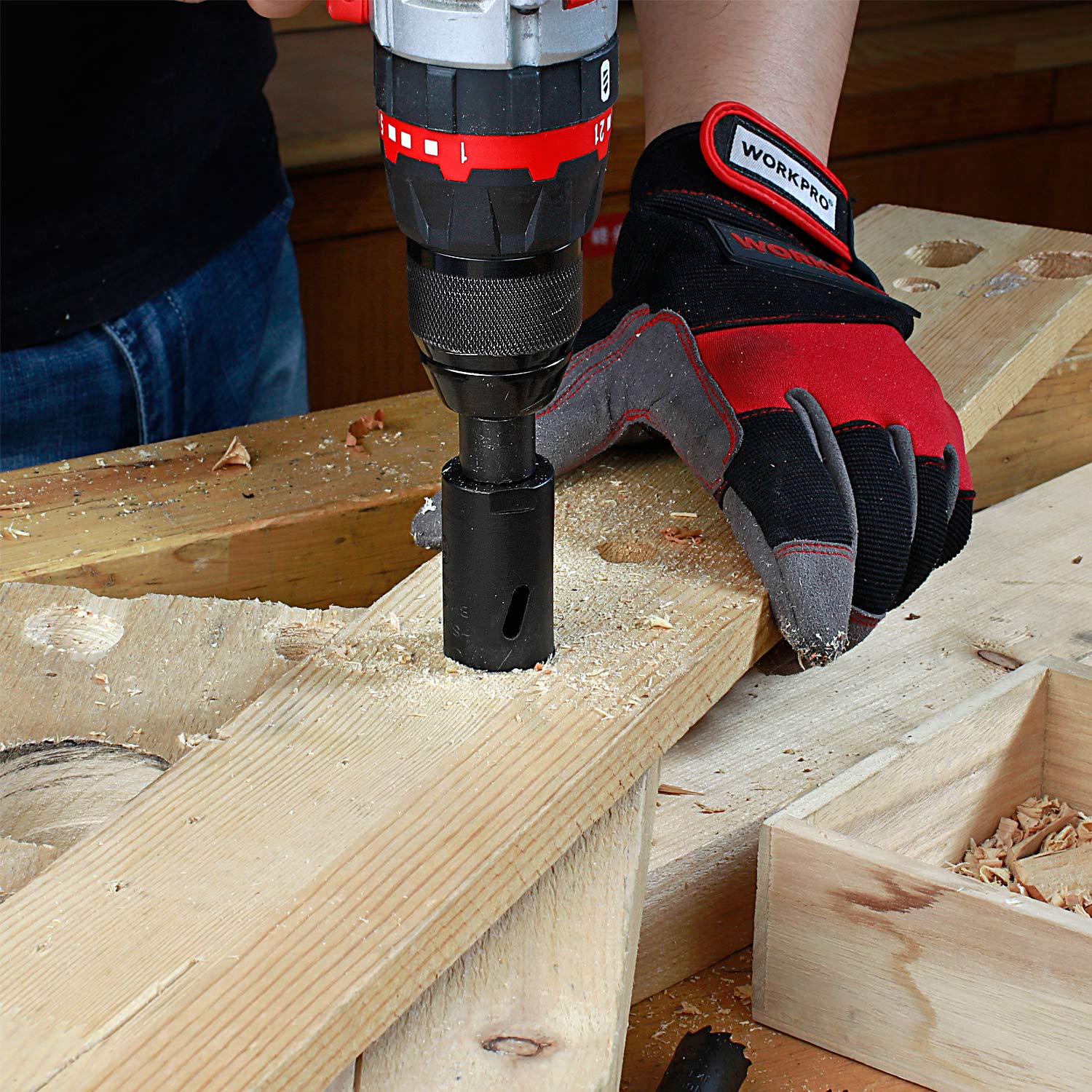 A person wearing gloves using a cordless drill with a WORKPRO hole saw to cut a hole in a wooden plank, producing wood chips.