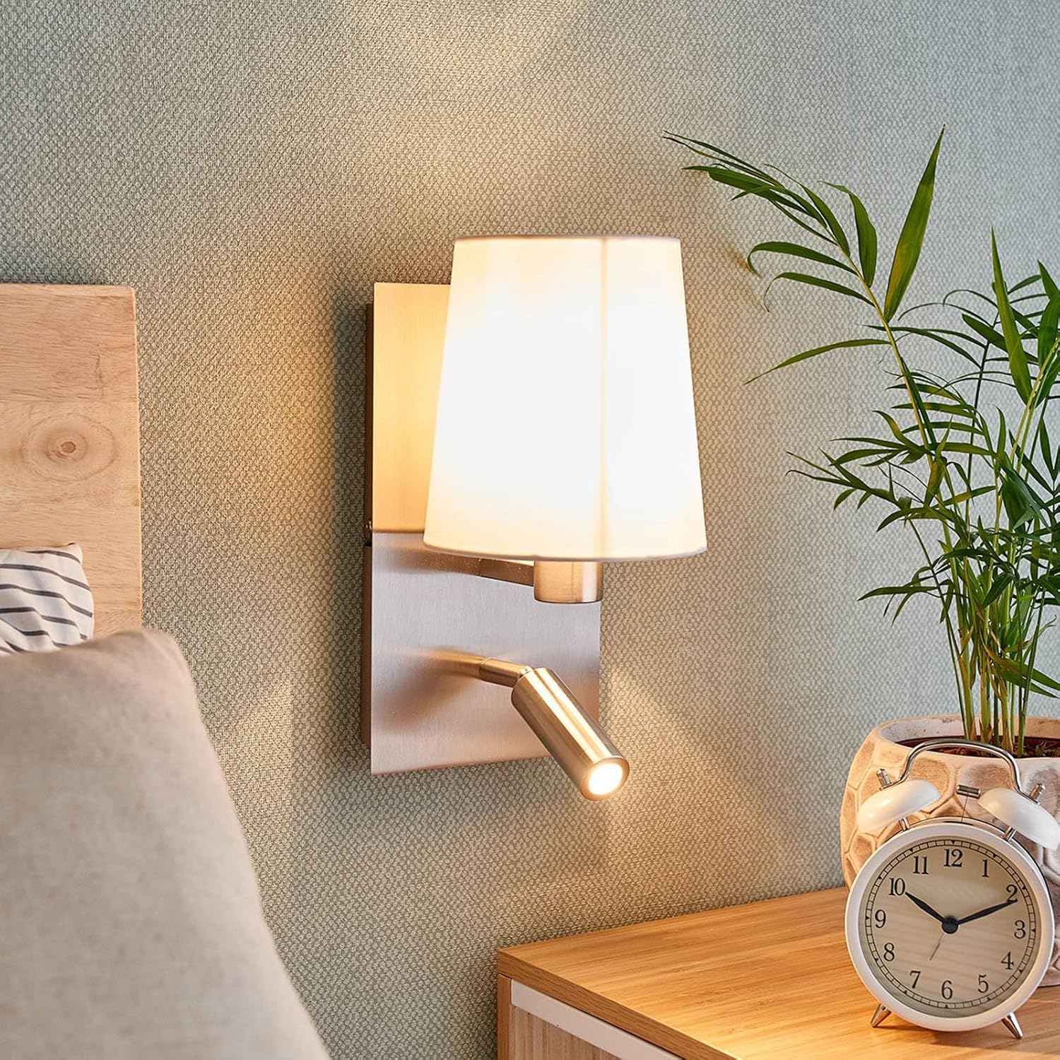 Lindby Aiden LED Wall Lamp installed in a bedroom setting, providing both ambient light from the main shade and focused light from the reading arm.