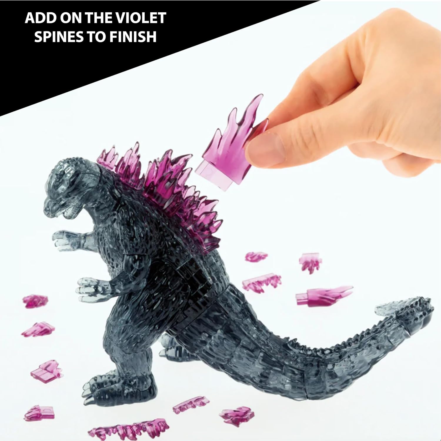 A hand placing a violet spine piece onto the back of a partially assembled translucent black Godzilla puzzle.