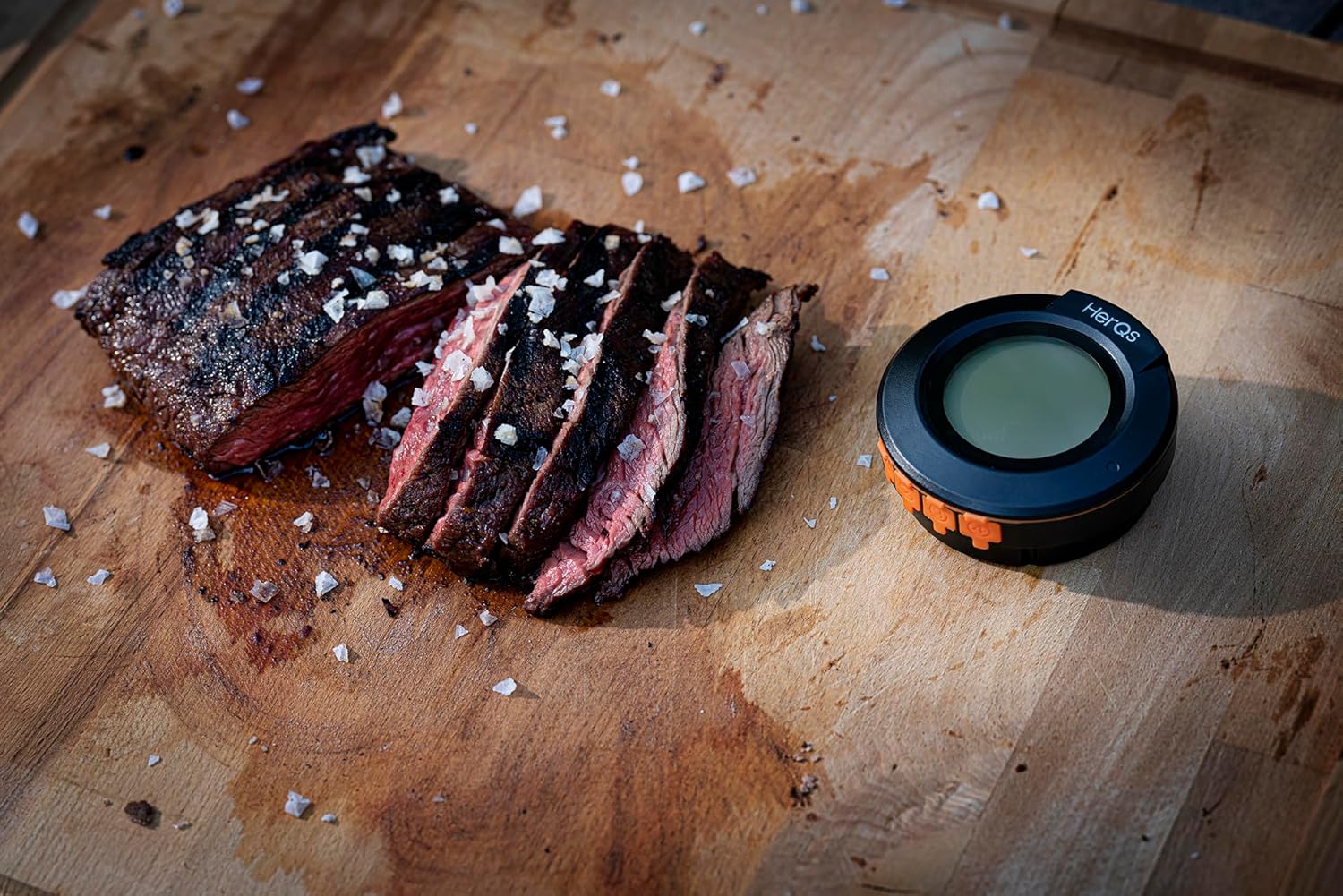HerQs thermometer placed next to perfectly sliced grilled steak on a wooden cutting board.
