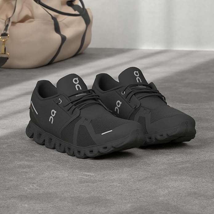 Pair of black On Men's Cloud 5 Running Shoes with mesh design and speed lacing, placed on a concrete floor next to a beige gym bag.