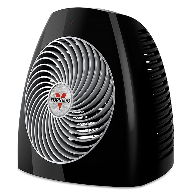 Vornado MVH 2025 whole room space heater in black with the signature Vornado vortex grille on the front, mechanical thermostat dial on top, carrying handle integrated into the chassis, sitting on a hardwood floor in a sunlit living room — the 2025 successor to the legendary VH200 lineage Wirecutter has recommended since 2018 and GoodHousekeeping named 2026 Best Overall
