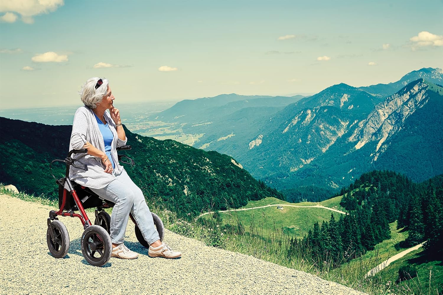 A senior woman sitting on the seat of the TAiMA XC Outdoor Rollator, enjoying a view of mountains and a lake.