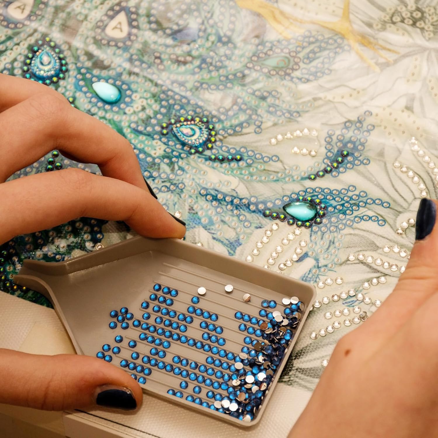 Hands using a diamond painting pen to pick up blue diamond beads from a gray tray, with a partially completed diamond painting canvas in the background.