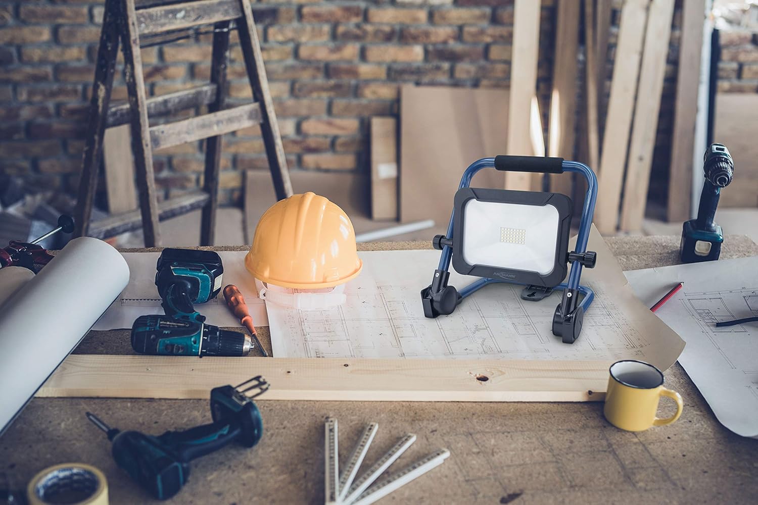 ANSMANN FL1600R 20W Cordless LED Work Light positioned on a workbench in a workshop setting, alongside tools and blueprints.