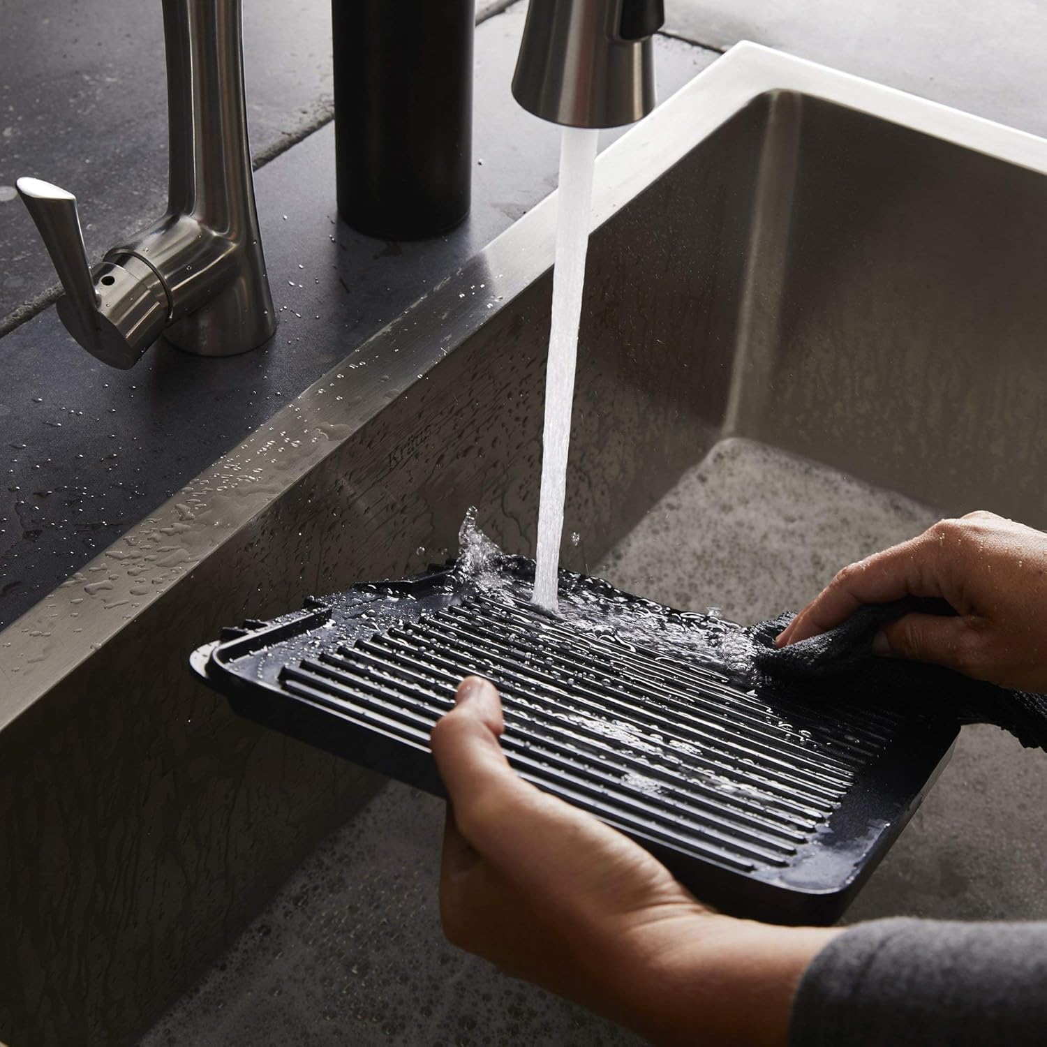 Hands washing a ribbed grill plate under running water in a kitchen sink.