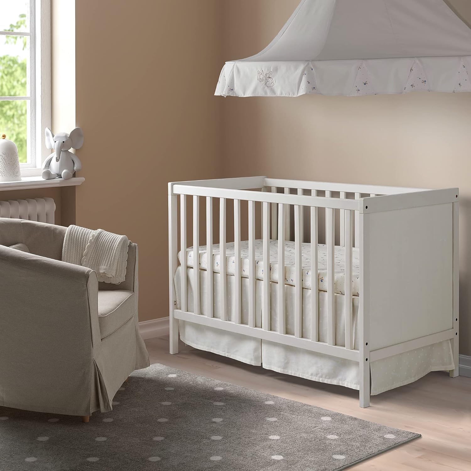 A white IKEA SUNDVIK cot with a mattress inside, set up in a nursery room with a chair and rug.